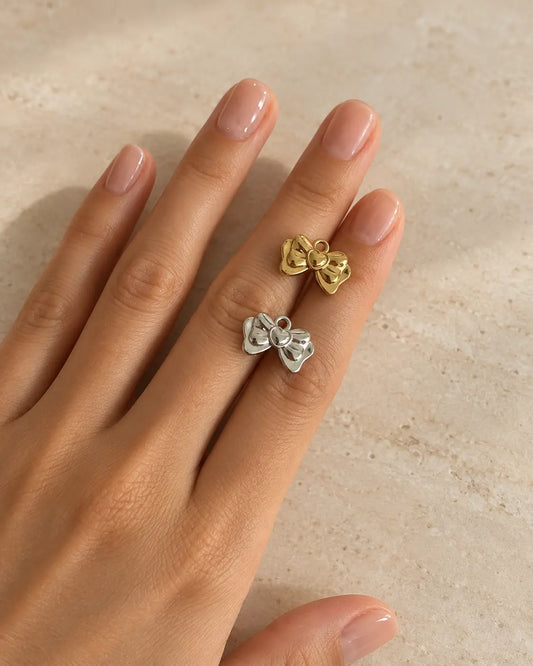 Goldener und silberfarbener Amelie Schleifen Charm mit Herzdetail auf einer Hand vor hellem Steinhintergrund