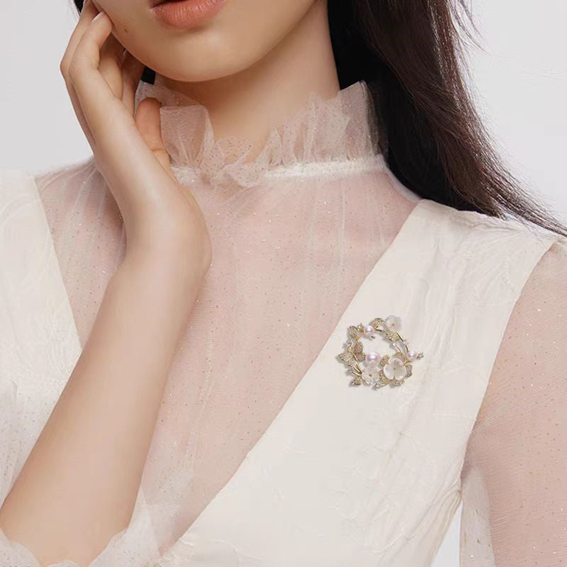 Elegante goldene Blumenkranz-Brosche mit Zirkoniablättern, geschnitzten Muschelblüten und rosa Süßwasserperlen – ideal für festliche Anlässe oder als Geschenk