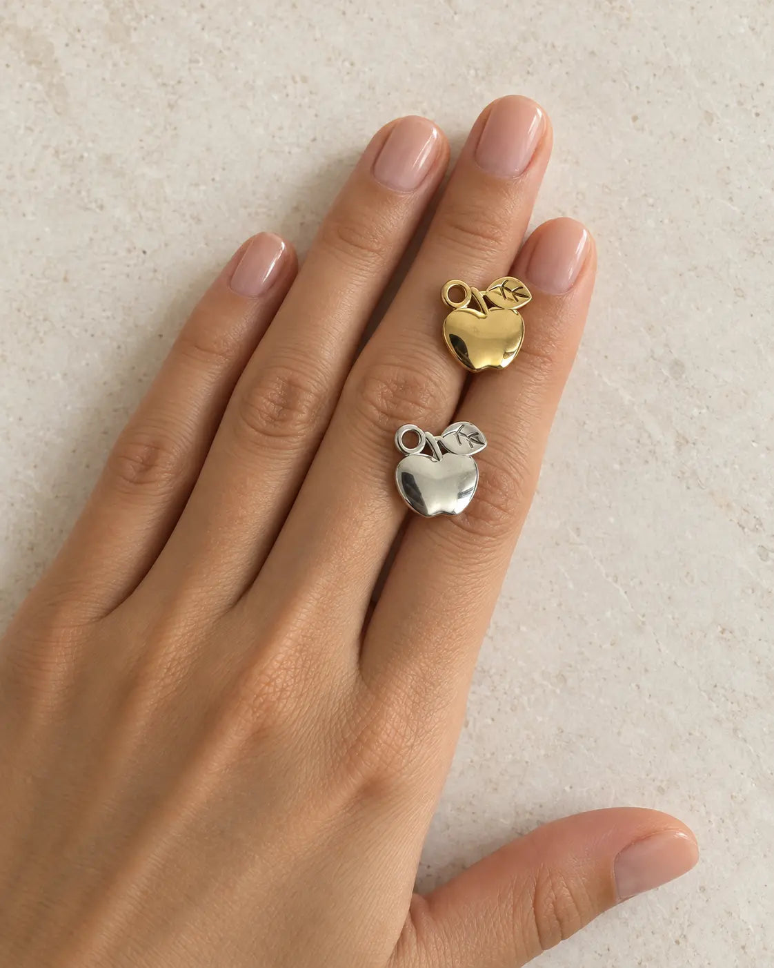 Goldener und silberfarbener Alma Apfel Charm auf einer Hand vor hellem Steinhintergrund