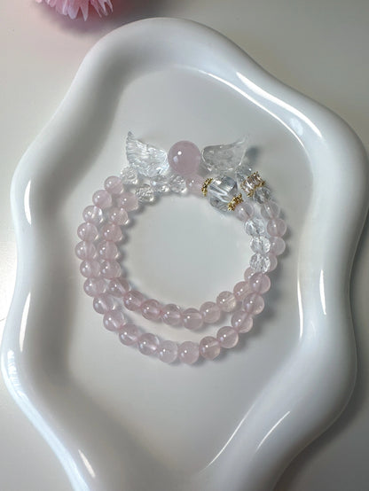 Doppellagiges Armband mit Rosenquarz, Bergkristall und Engelsflügel-Anhänger auf weißem Teller – zart und spirituell