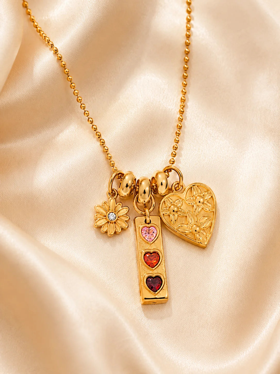 Goldene Kugelkette aus 18K vergoldetem Titanstahl mit drei verschiedenen Anhängern: einer Blume, einem länglichen Anhänger mit drei bunten Herzsteinen und einem geprägten Herz.