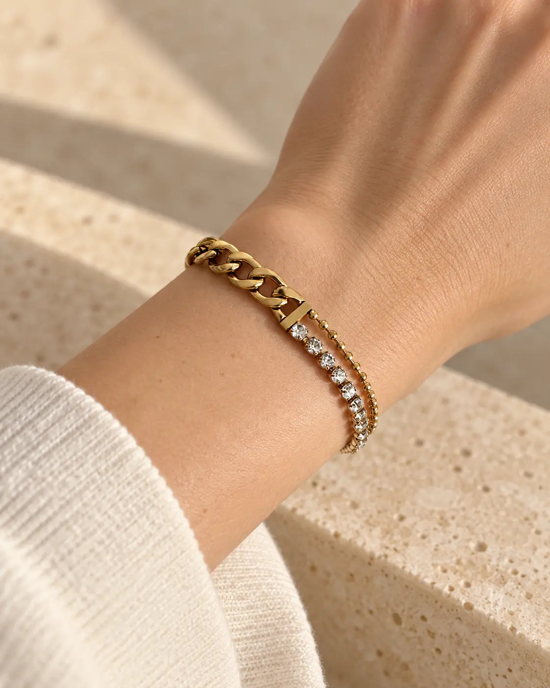 Goldenes Nerina Zirkonia Armband am Handgelenk mit hellem Strickärmel vor beigem Steinhintergrund