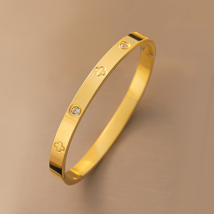 Vergoldeter Armreif mit Kleeblatt-Gravur und funkelnden Zirkonia-Steinen auf beigem Hintergrund