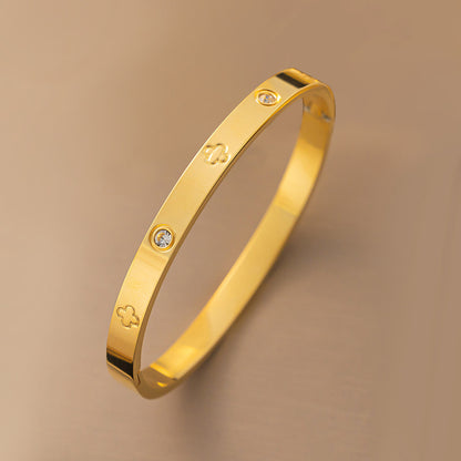 Vergoldeter Armreif mit Kleeblatt-Gravur und funkelnden Zirkonia-Steinen auf beigem Hintergrund