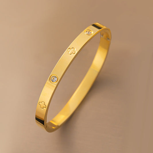 Vergoldeter Armreif mit Kleeblatt-Gravur und funkelnden Zirkonia-Steinen auf beigem Hintergrund