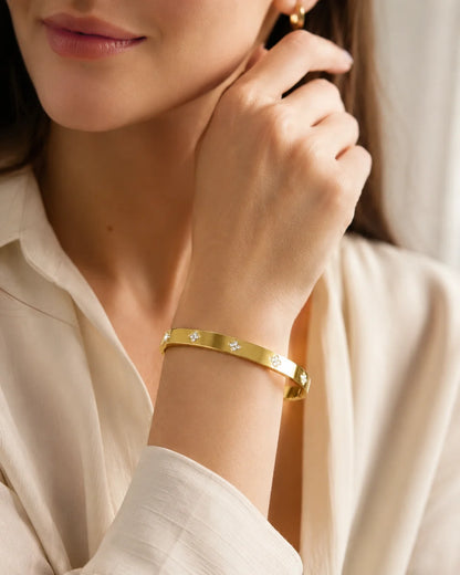 Gold-tone cubic zirconia clover bangle with small floral motifs, worn on the wrist beside a pale blouse.